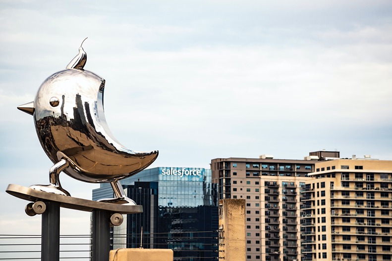 The shiny silver sculpture of a bird on a skateboard in front of the Dallas skyline