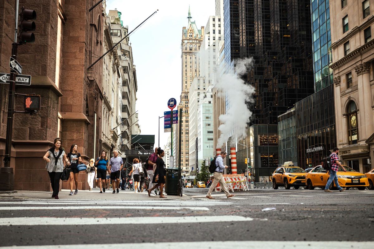 Pedestrians crossing a Manhattan street with yellow taxis, steam rising from a vent and skyscrapers behind