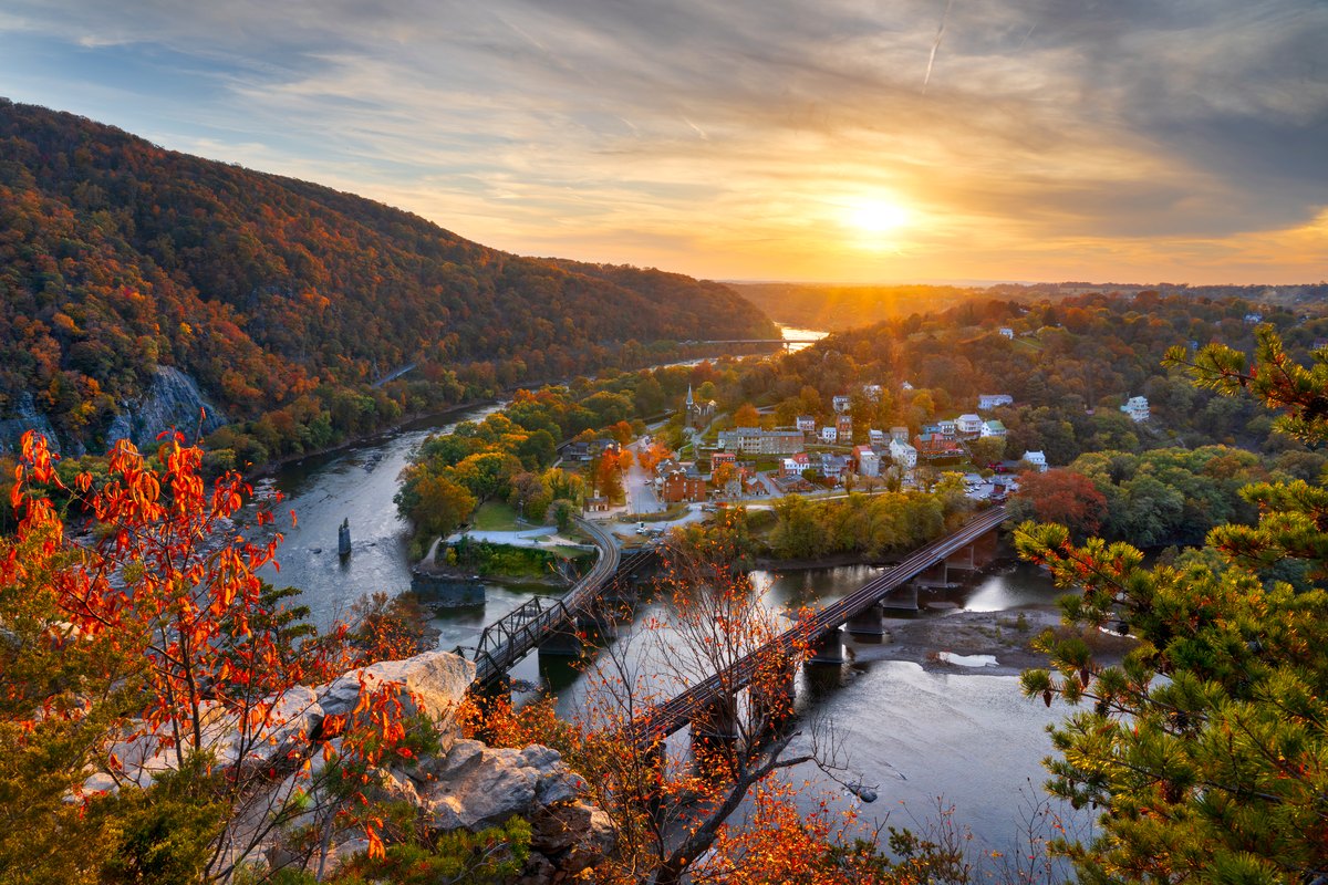 The town of Harpers Ferry at the meeting of the Potomac and the Shenandoah at sunrise, a railway bridge crossing the water below wooded ridges