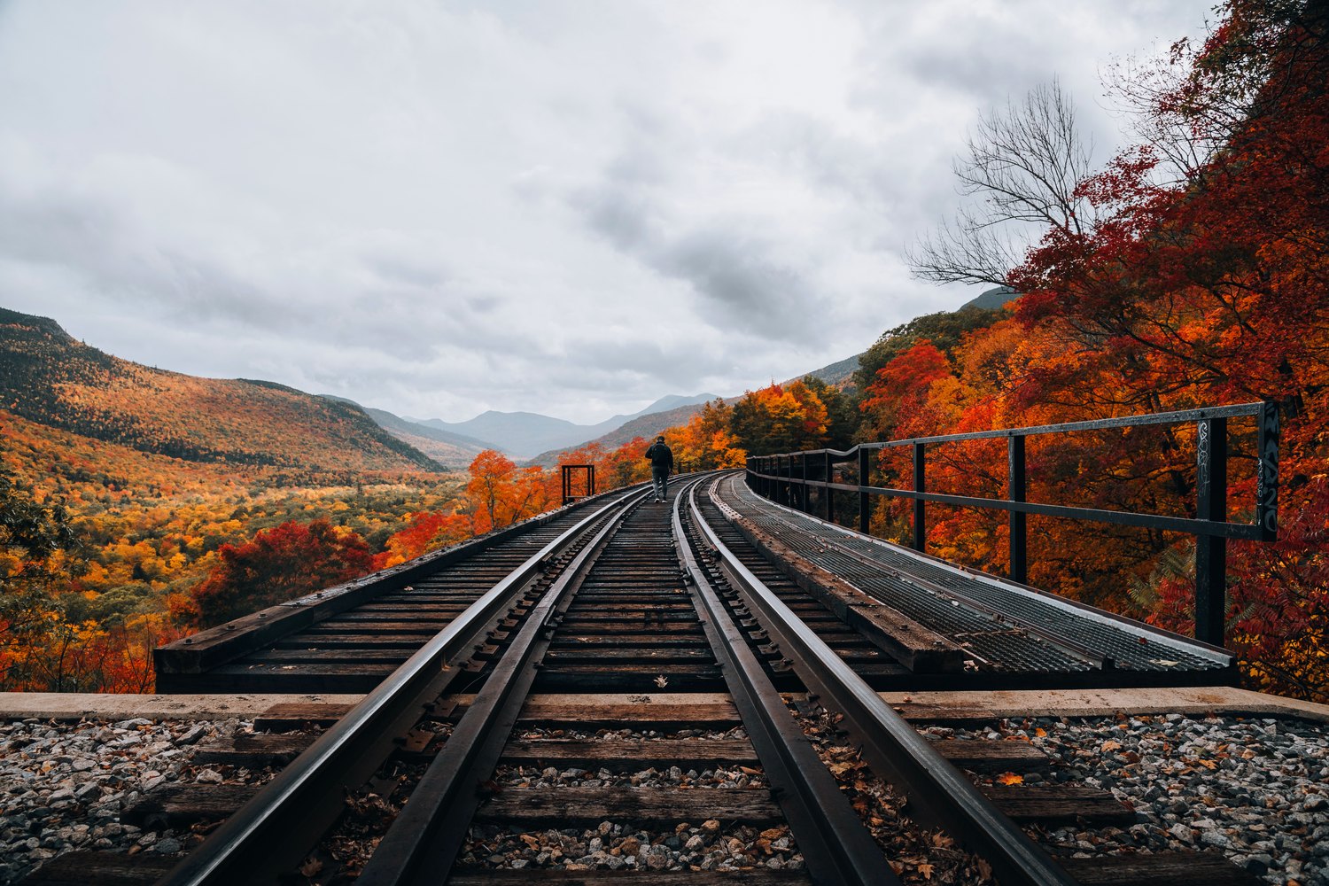 Railway track curving across a trestle through a New Hampshire notch, red maples crowding the right-hand side under a broad cloudy sky