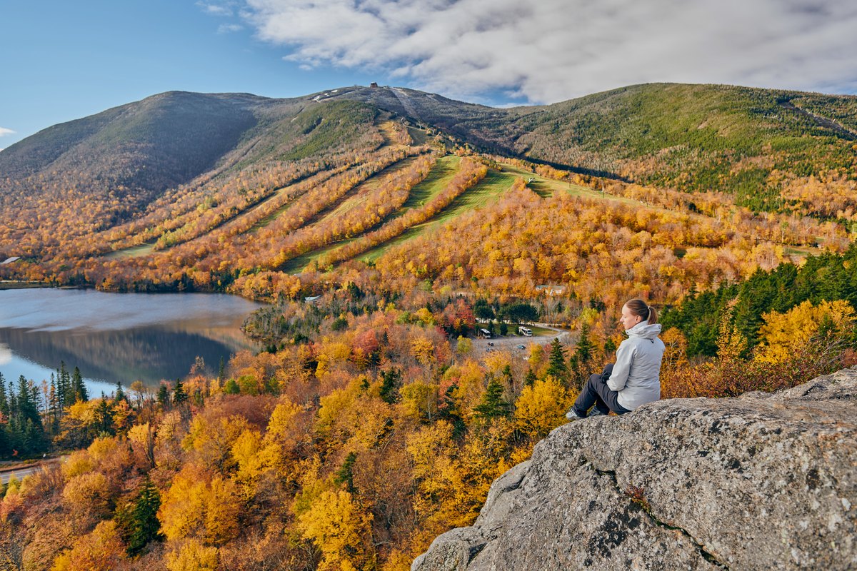 A hiker sitting on a rock ledge above a mountain lake, the far slope covered in orange and gold hardwood
