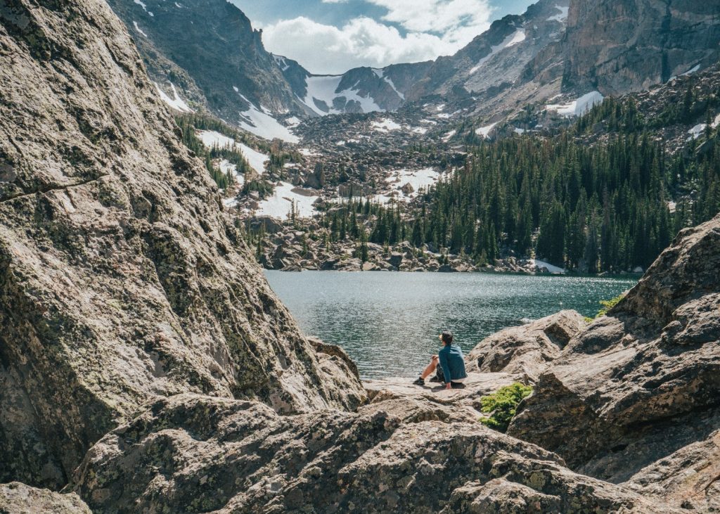 A hiker in Estes Park sits on boulders overlooking an alpine lake and snowy mountains