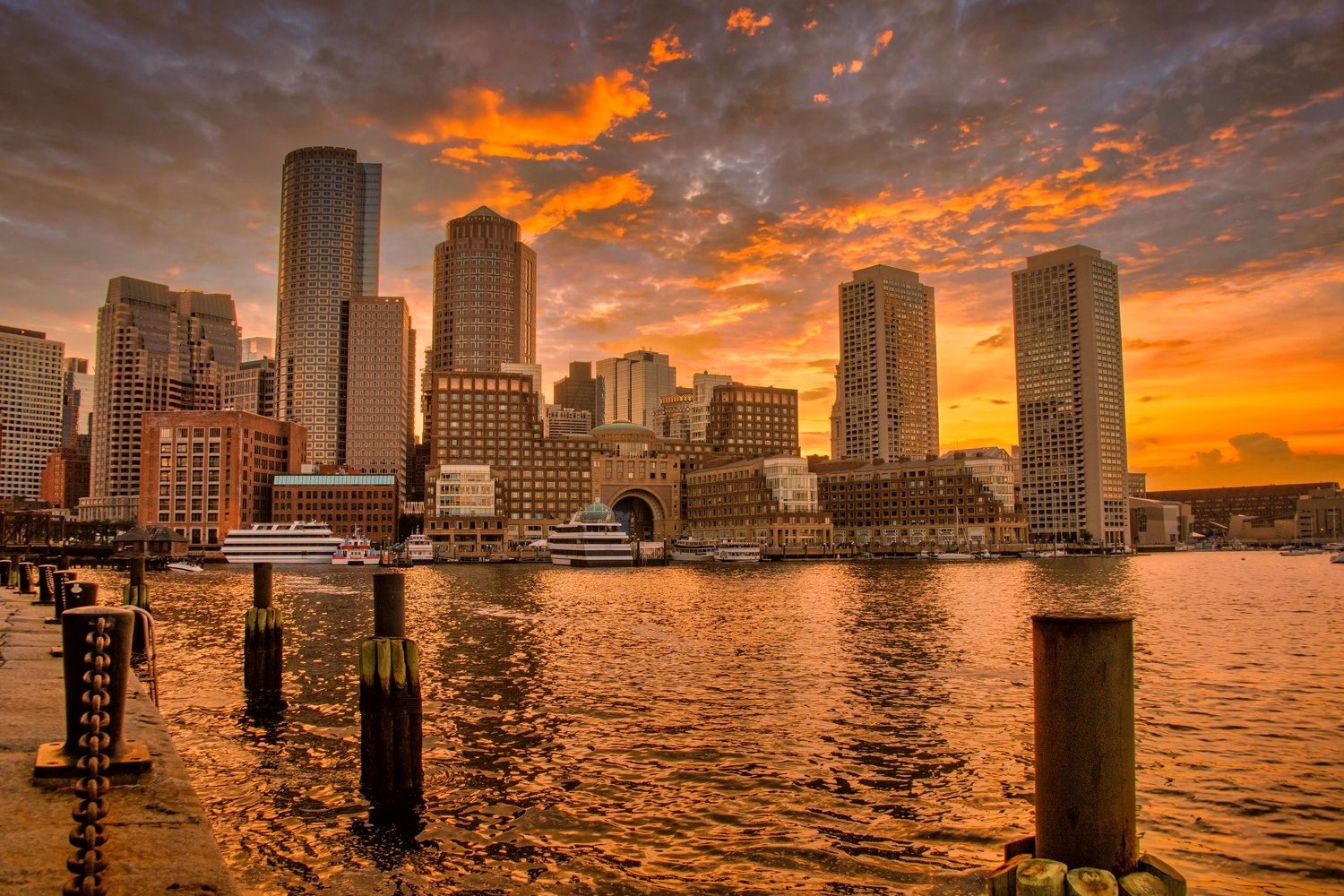 The Boston skyline at dusk across the harbor, golden sunset light behind the towers and wooden pier posts in the foreground