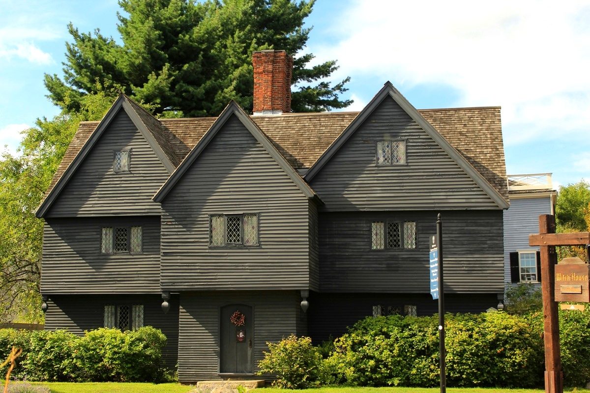 The Witch House in Salem, dark clapboard and steep gables above a green hedge, the museum sign on its corner post