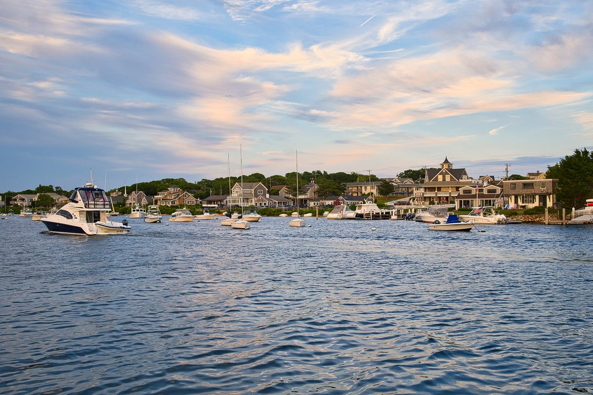 Boats moored in Falmouth Inner Harbor on Cape Cod under an evening sky, with shingled waterfront houses behind them