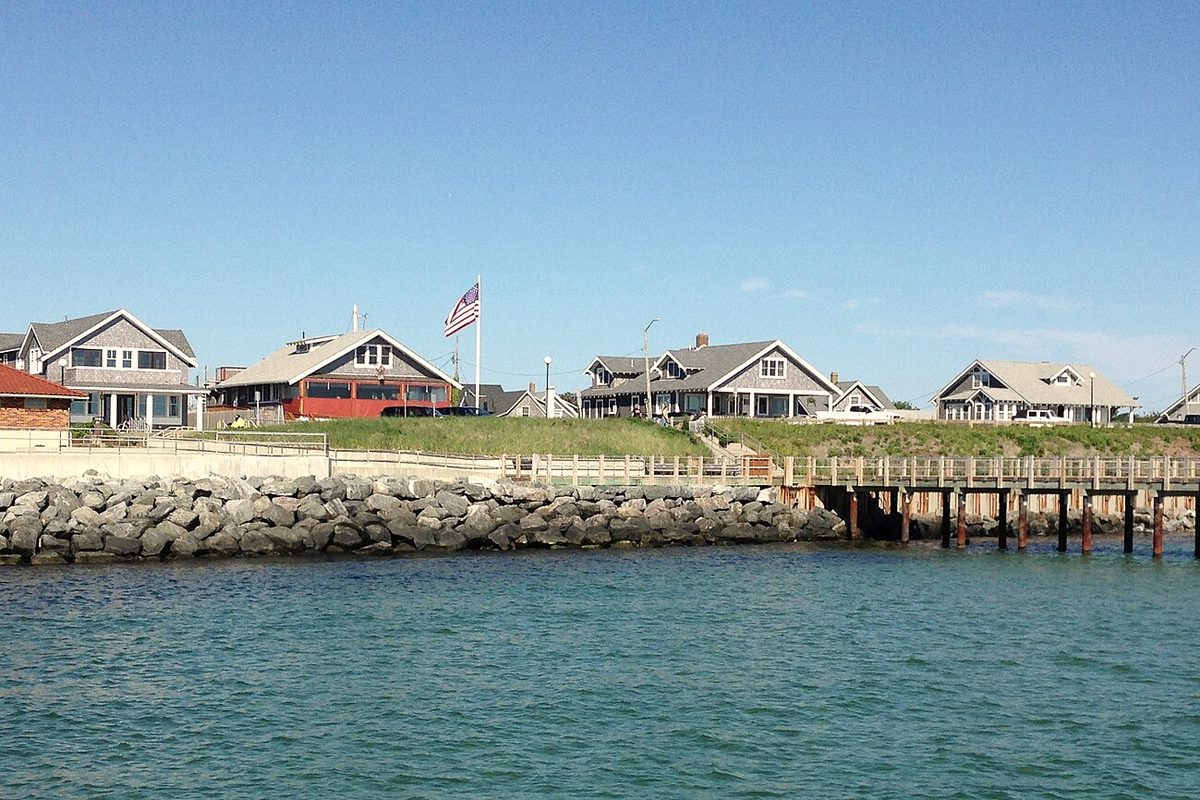 The Oak Bluffs waterfront on Martha’s Vineyard seen from the ferry, with shingled houses on a grassy bluff above a stone seawall and a wooden pier