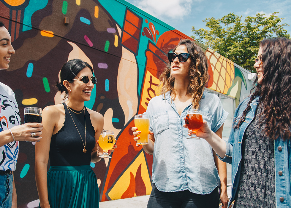 Friends drink beer in the sun in front of a wall of graffiti.
