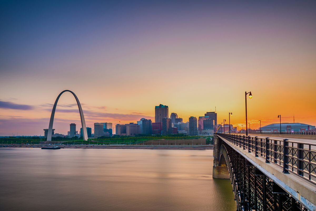 The Gateway Arch and the St. Louis skyline at sunset from the Eads Bridge walkway, the railing running toward downtown