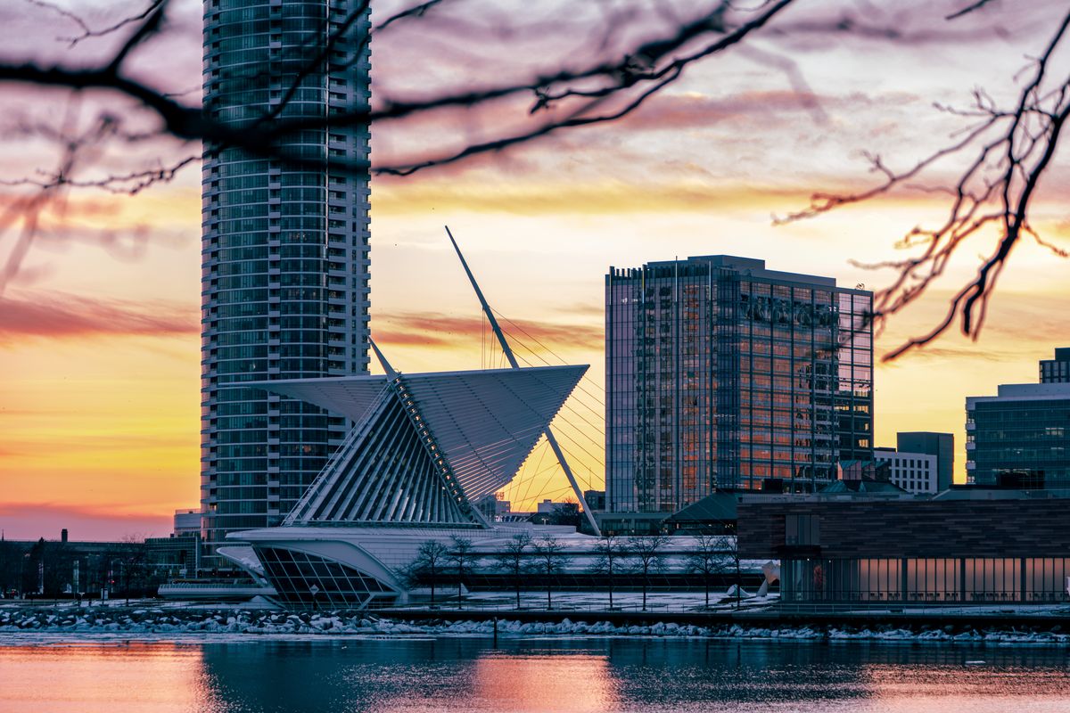 The Milwaukee skyline and the winged roof of the Milwaukee Art Museum at sunset, seen through bare branches across the water