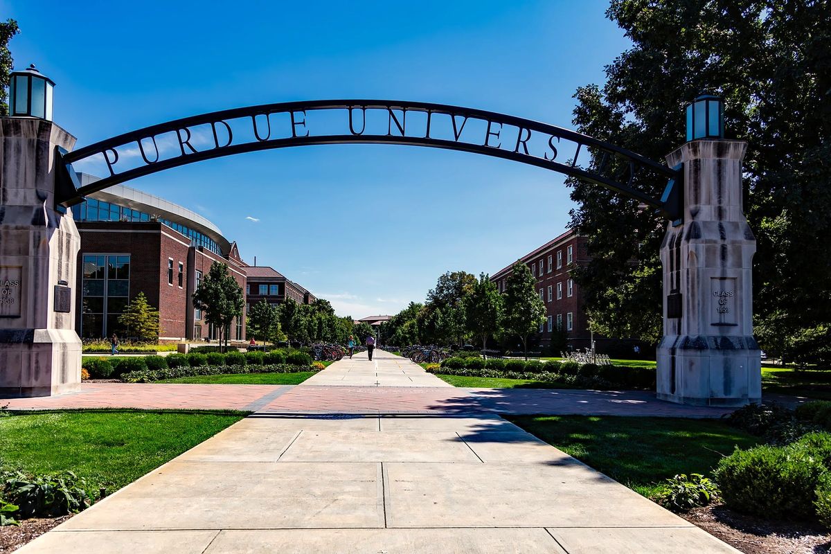 The Purdue University gateway arch over a long campus walkway on a clear summer day, a lone student walking beneath it