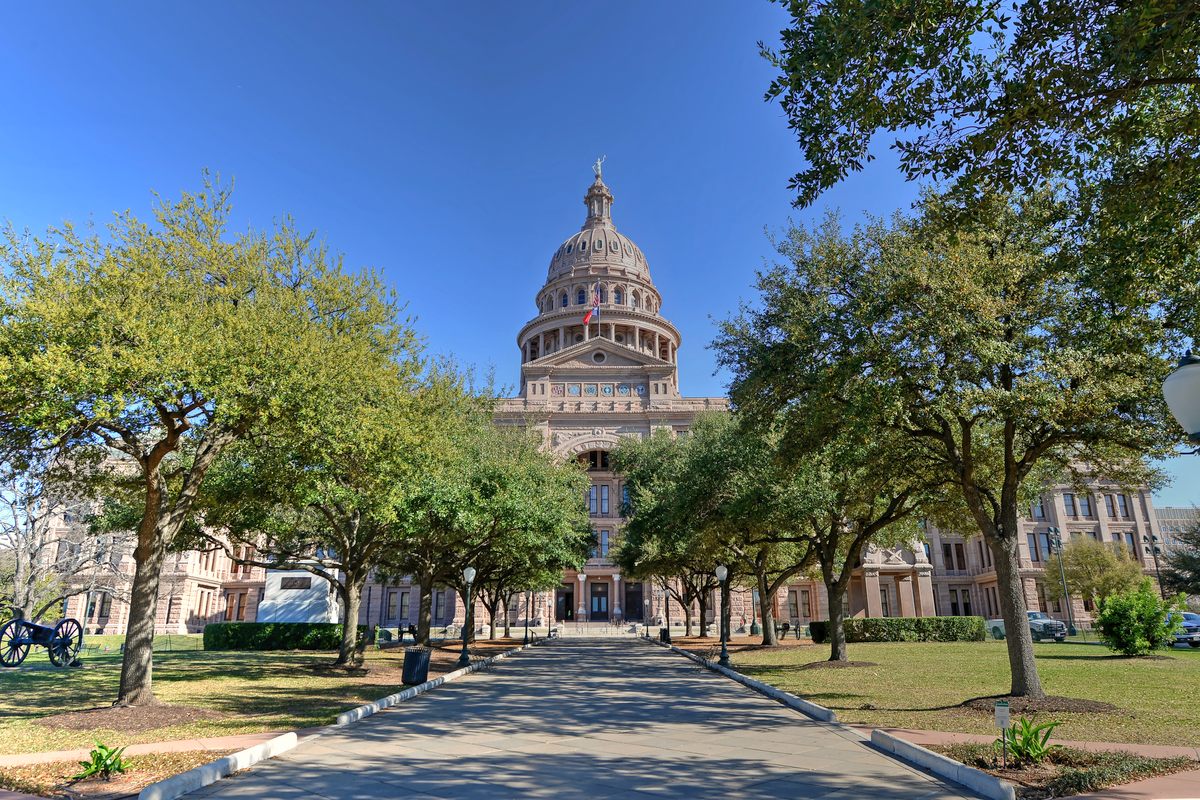The Texas State Capitol in Austin, its pink granite front and tall dome under a clear blue sky, seen down a tree-lined walkway across the capitol lawn