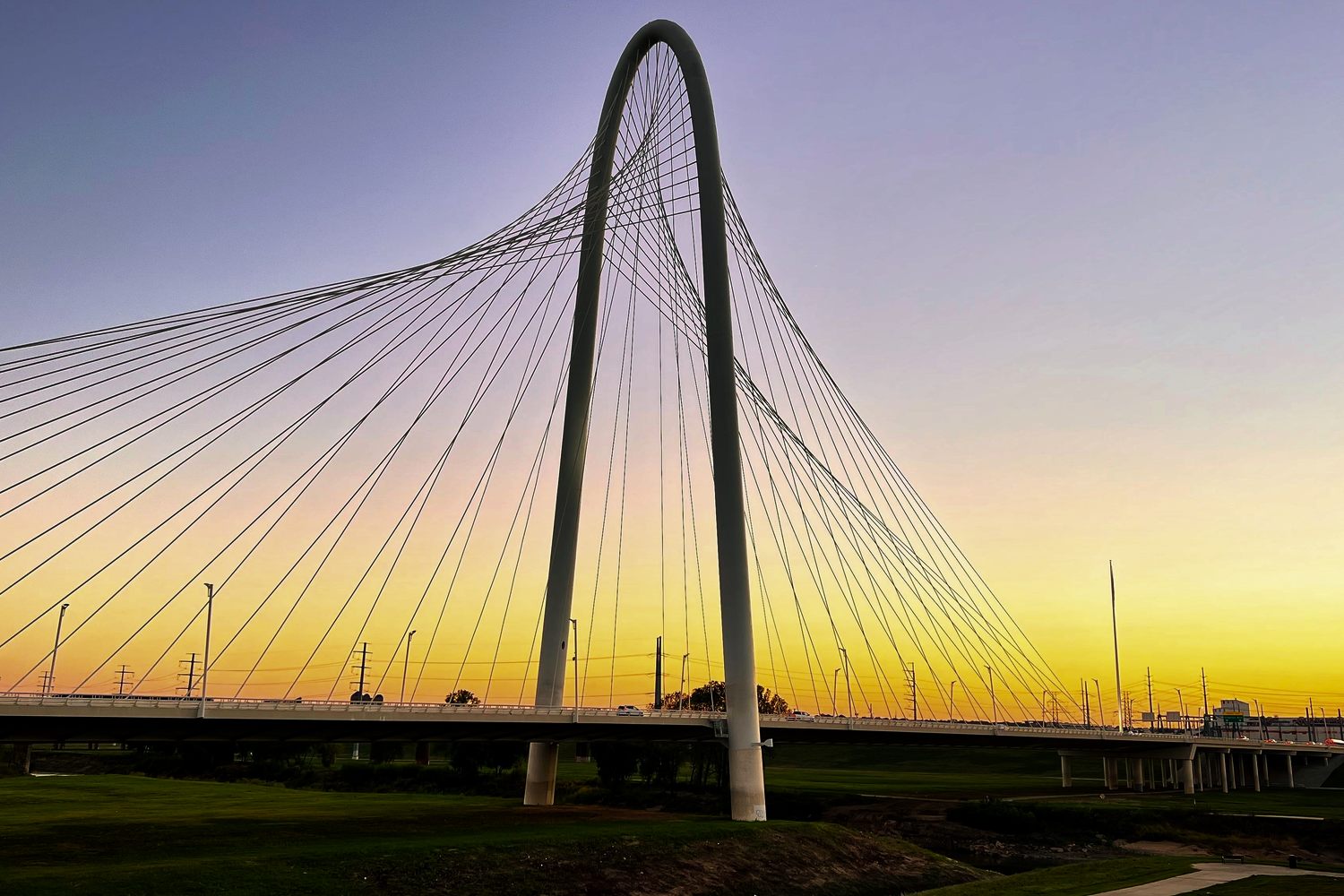 The white arch of the Margaret Hunt Hill Bridge in Dallas against a yellow and violet sunset, its cables fanning down to the roadway above the grassy Trinity River floodplain