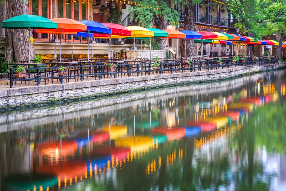 Rows of red, orange, yellow, green and blue umbrellas over café tables on the San Antonio River Walk, their colors reflected in the still water of the river below a stone wall