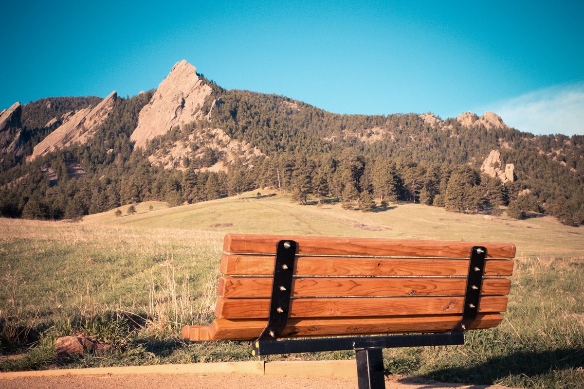 A wooden bench on the grassy meadow at Chautauqua in Boulder, facing the tilted sandstone slabs of the Flatirons above a pine-covered slope under a clear blue sky