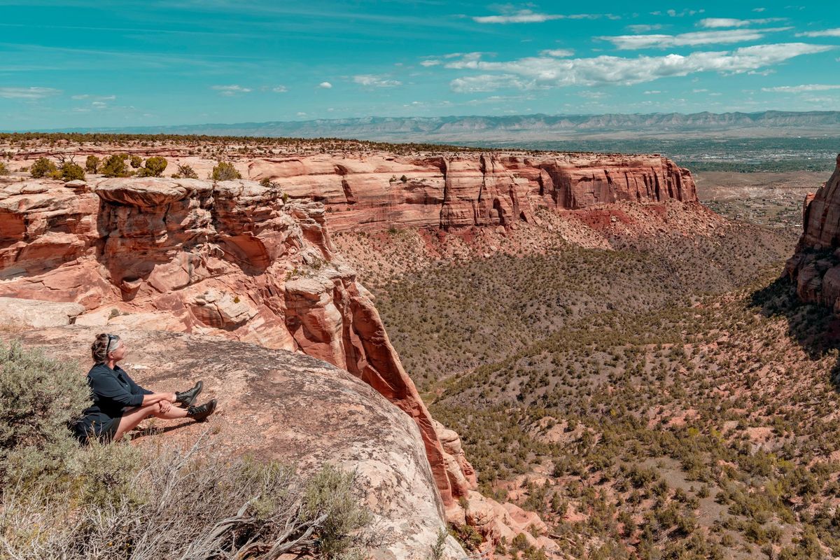 A visitor sits on a sandstone ledge at Colorado National Monument, looking across a red-rock canyon and green scrub toward the Grand Valley under a blue sky with thin clouds