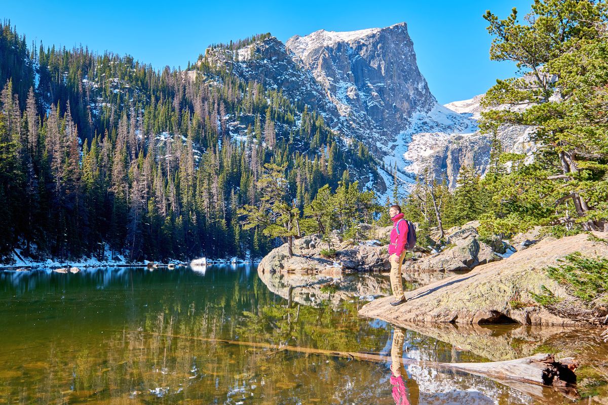 A hiker in a pink jacket stands on a rock ledge at Dream Lake in Rocky Mountain National Park, with snowy cliffs and dark pines reflected in the still water