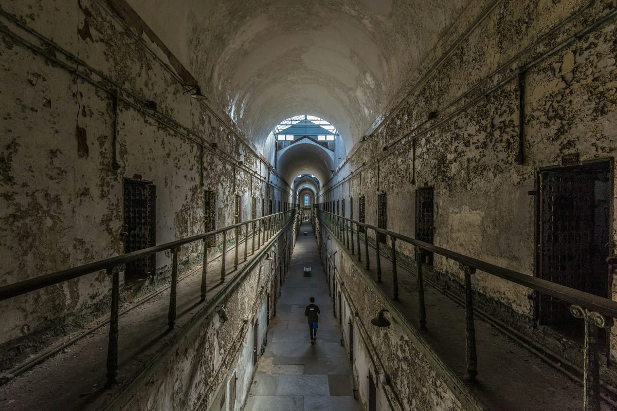 A long, crumbling cellblock corridor at Eastern State Penitentiary in Philadelphia, lit by skylights, with a lone visitor walking away