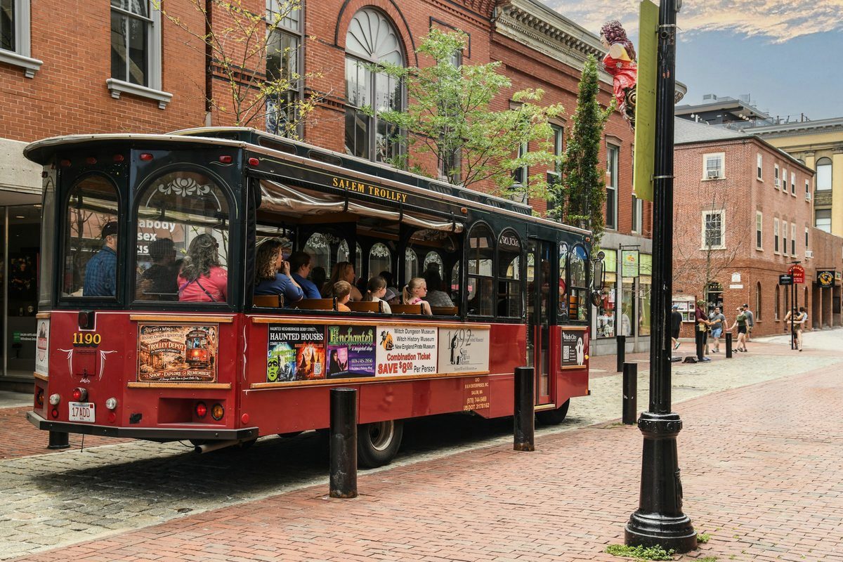 The red Salem Trolley full of travelers rolling along a brick street in downtown Salem, Massachusetts, past Halloween-decorated storefronts