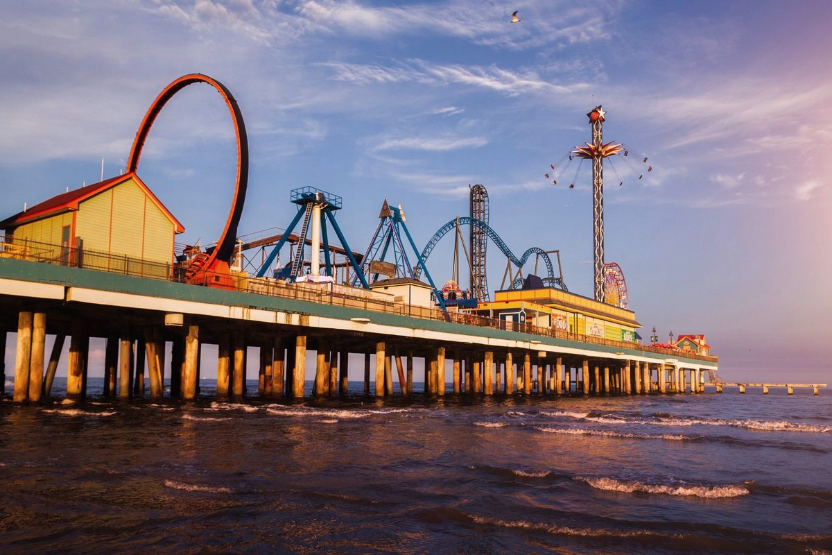 The Galveston Island Pleasure Pier at golden hour, its red loop ride, blue roller coaster and star-topped swing tower standing on pilings above the Gulf surf