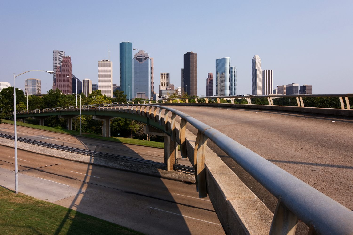 The downtown Houston skyline on a clear day, glass and granite towers rising above the trees beyond a curving concrete freeway overpass with a steel railing