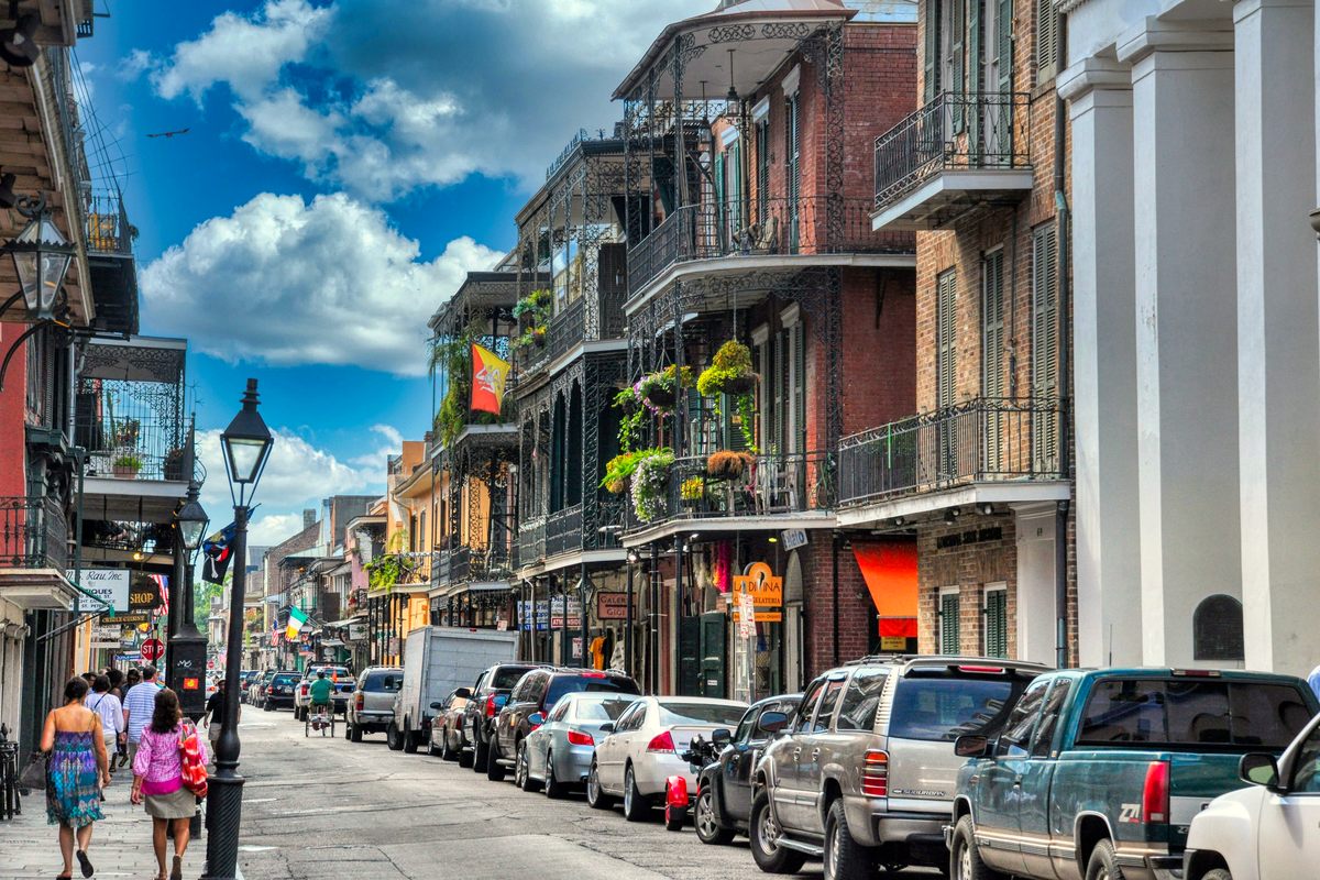 A French Quarter street in New Orleans, brick buildings with wrought-iron balconies hung with ferns and flowers, cars parked at the curb and people on the sidewalk under a blue sky