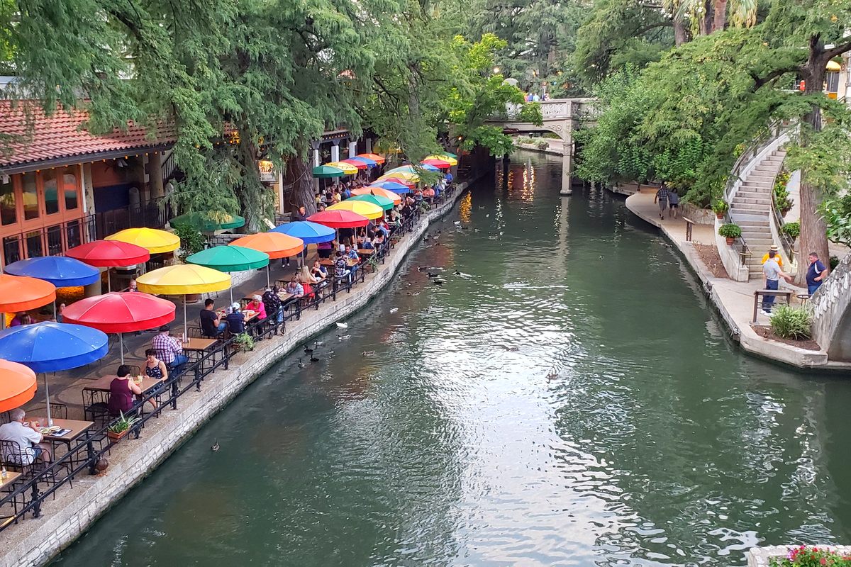 Diners under red, blue, yellow and orange umbrellas along the San Antonio River Walk, the green river curving past under cypress trees toward a stone footbridge