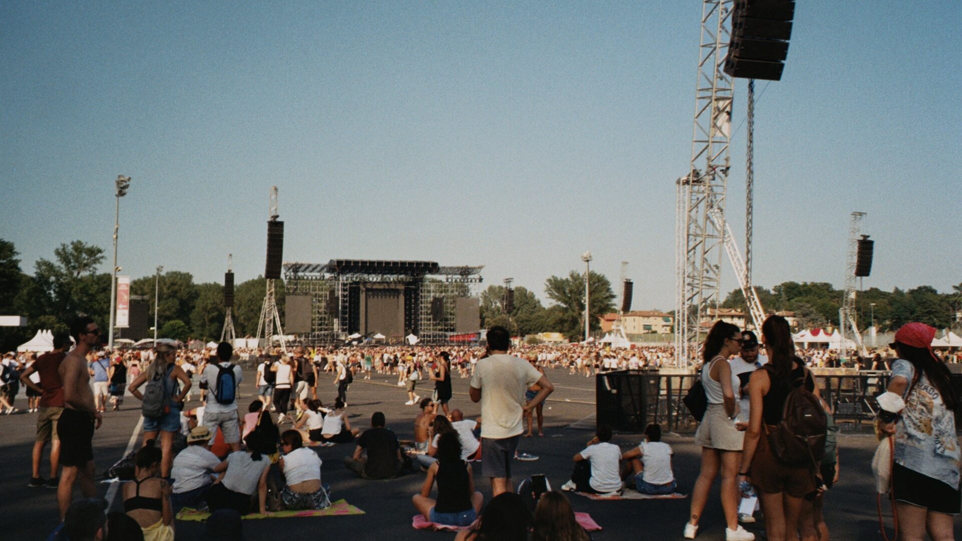 Festival crowd standing and sitting on open ground under a clear sky, with the stage far in the distance behind speaker towers