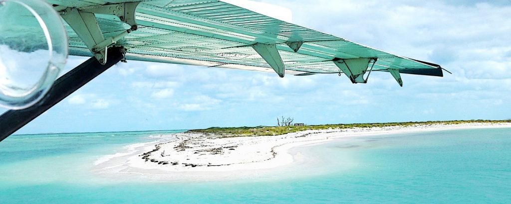The tip of an airplane wing visible as it flies over a strip of sand in the bright teal waters of the Florida Keys