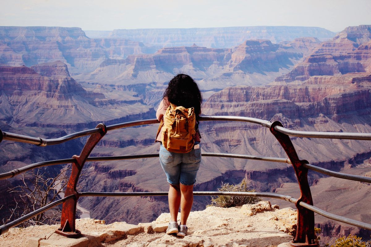 A traveler with a tan backpack leans on the railing at a South Rim overlook, looking out across the Grand Canyon’s layered red and violet buttes as they fade into haze toward the far rim