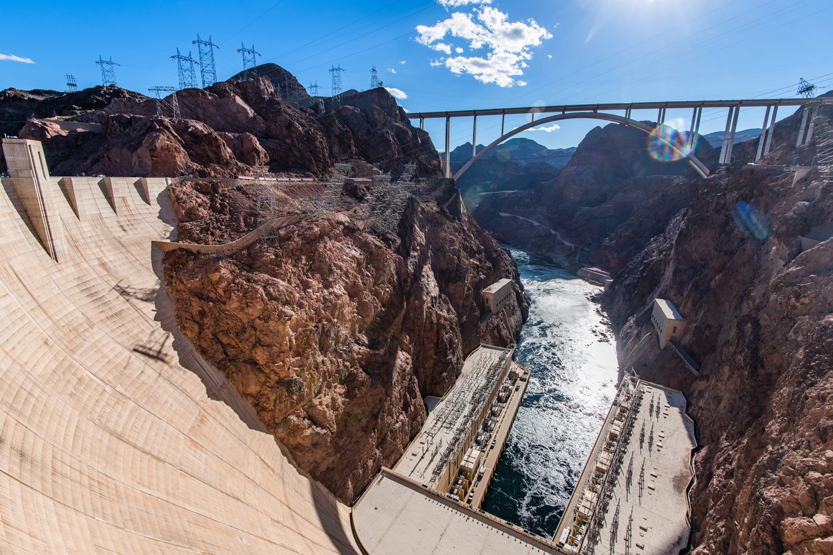 Looking down from the top of Hoover Dam’s curved concrete face into the canyon, with the powerhouses and the Colorado River below and the arch of the Mike O’Callaghan-Pat Tillman Memorial Bridge spanning the gorge against a blue sky