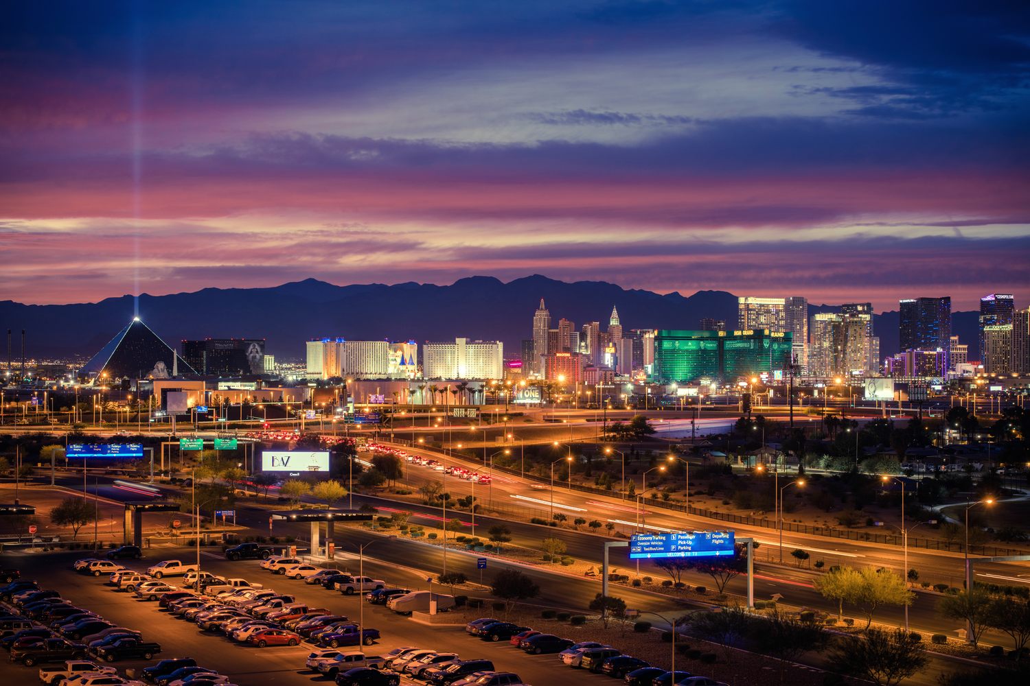 The Las Vegas Strip at dusk under a pink and violet sunset sky, the Luxor pyramid sending its light beam up at the left, the lit towers of the south Strip glowing gold and green across the middle, and a ridge of dark desert mountains along the horizon
