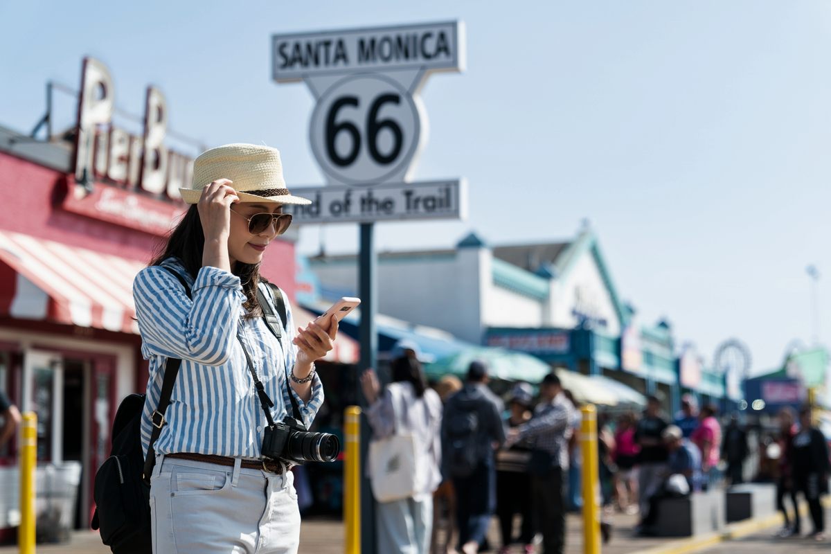A traveler in a straw hat and sunglasses checks her phone under the Santa Monica Route 66 End of the Trail sign on the pier, a camera slung at her side and the pier’s shops and crowds behind her