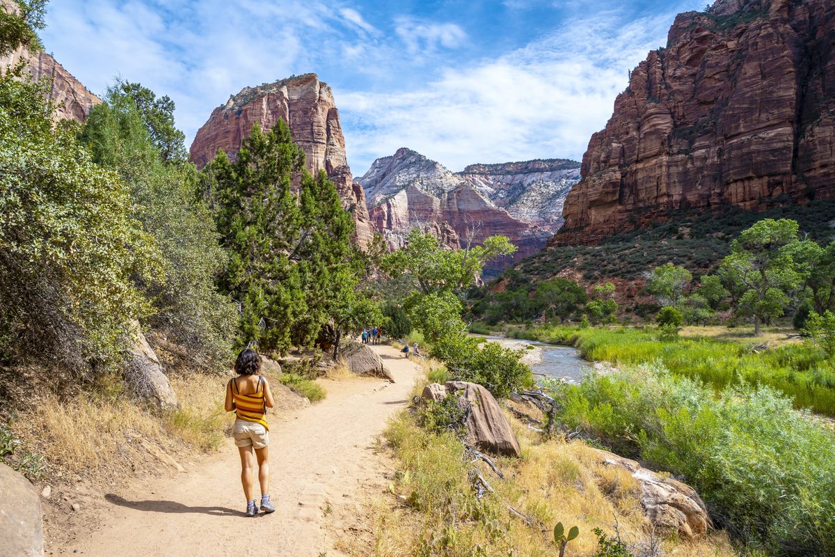 A hiker in a striped top walks a sandy trail beside the Virgin River in Zion Canyon, past cottonwoods and green brush, with red and white sandstone cliffs rising on both sides under a blue sky