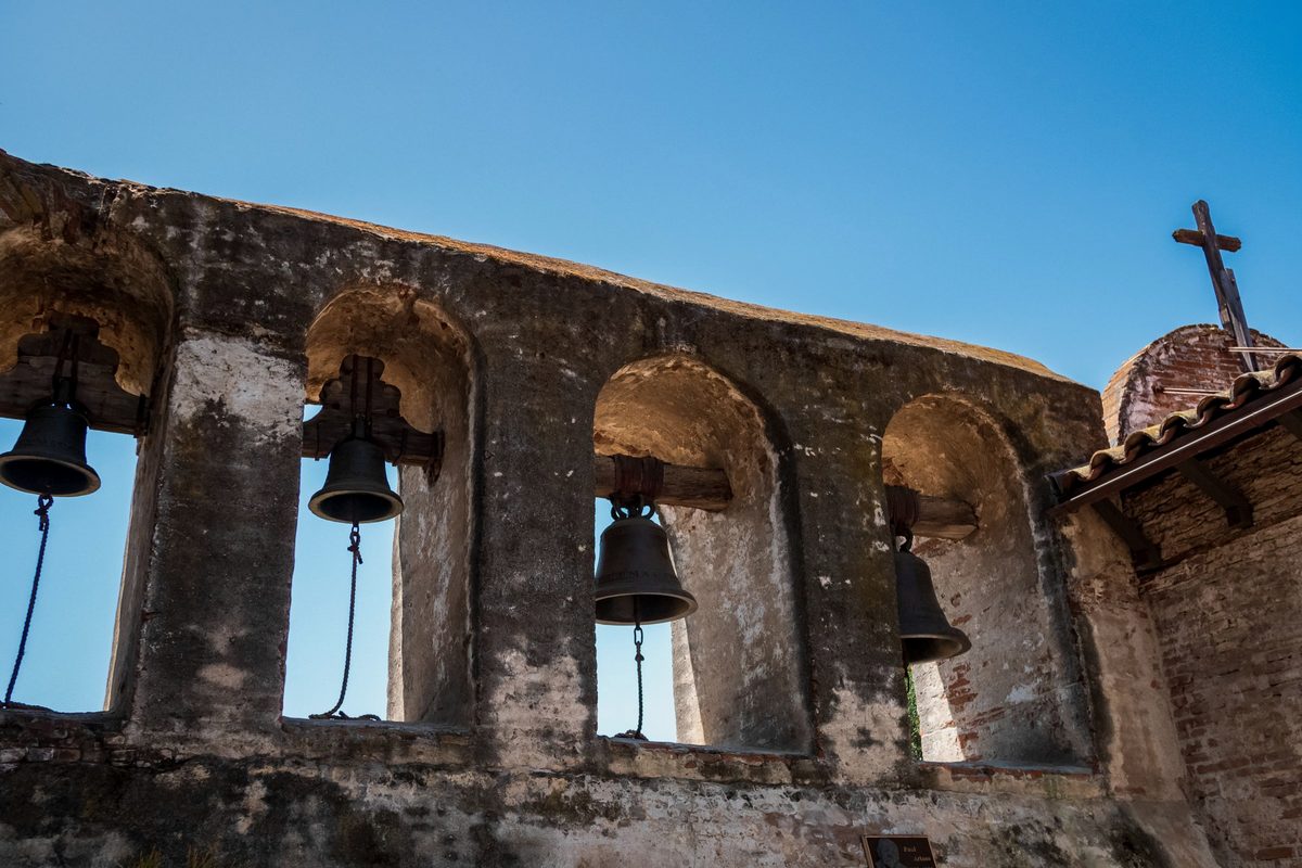 The bells of Mission San Juan Capistrano hanging in their weathered stone wall, seen from below against a clear sky