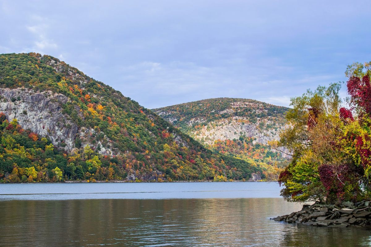 Autumn-colored hills of the Hudson Highlands meeting the river near Cold Spring, New York, under a soft blue sky