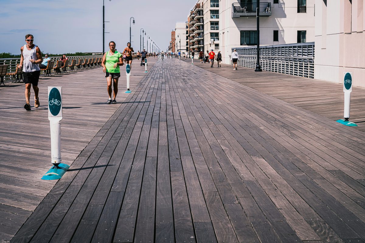 Joggers and walkers on the wide wooden boardwalk at Long Beach, New York, with oceanfront apartment buildings along one side on a sunny day