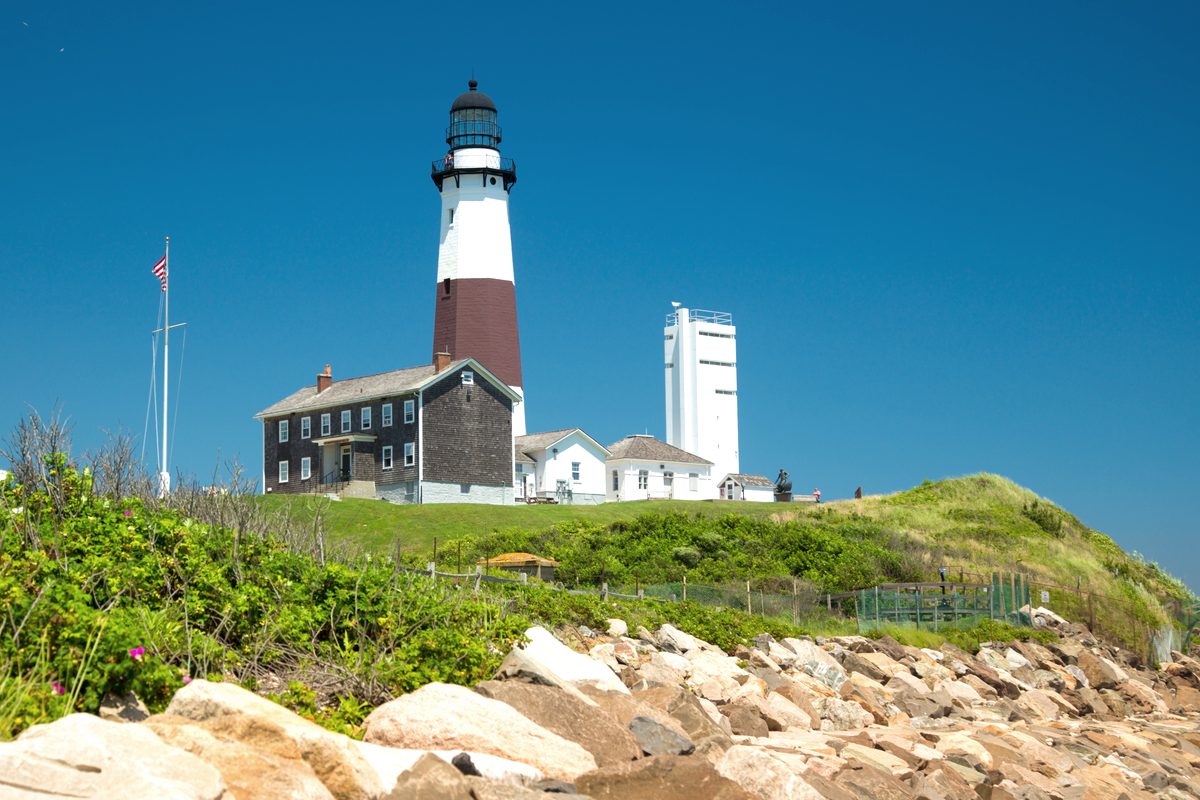 The white Montauk Point Lighthouse with its brown band, standing on a grassy bluff above the rocks at the eastern tip of Long Island