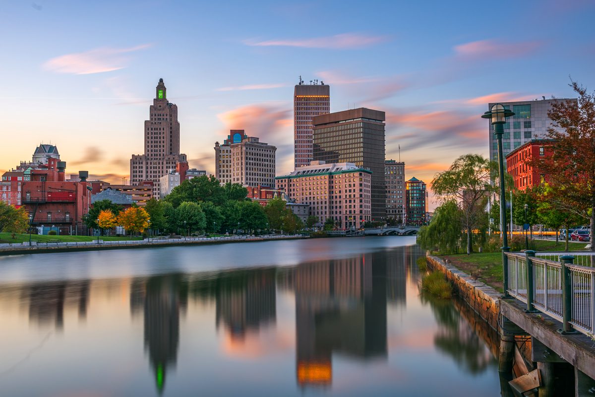 Downtown Providence reflected in the river at sunset, with trees and a railed riverside walkway in the foreground