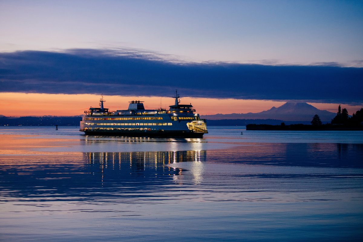 A Washington State ferry crossing Puget Sound off Bainbridge Island at dusk, its lit decks reflected in calm water, with Mount Rainier on the horizon under a band of orange sky
