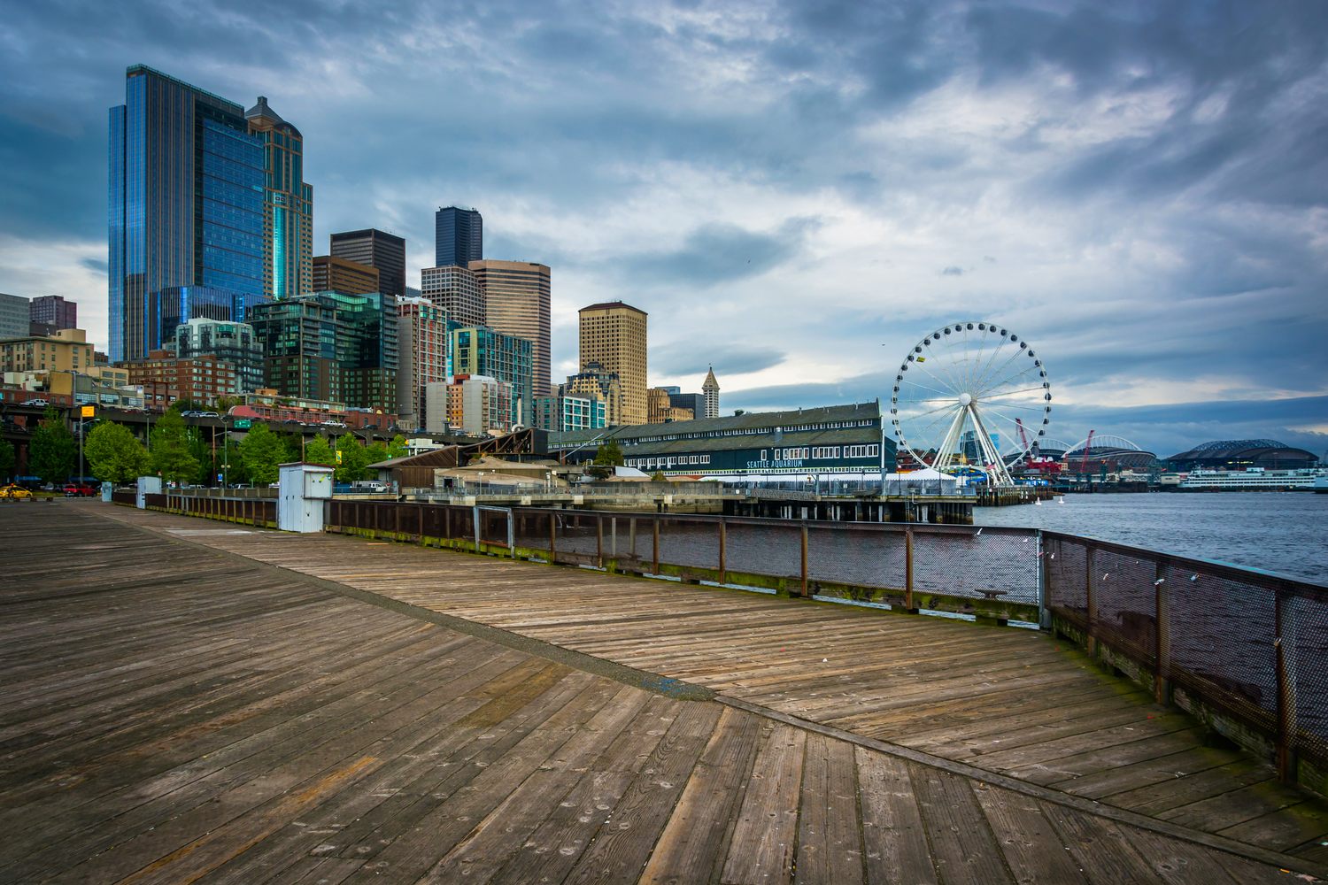 The downtown Seattle skyline under a heavy grey sky, seen from the weathered wooden deck of Piers 62 and 63, with the Seattle Aquarium, the Great Wheel and the stadiums along Elliott Bay to the right