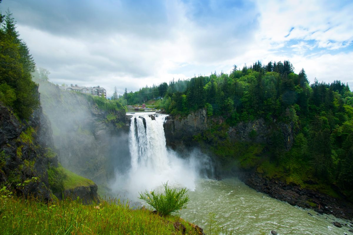 Snoqualmie Falls pouring into a misty gorge between forested cliffs, the Salish Lodge on the clifftop at upper left and the green river running out below