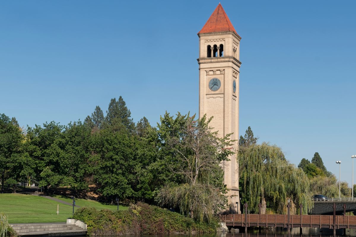 The brick clock tower in Spokane’s Riverfront Park, its red spire above willows and pines on a clear day, with a wooden footbridge over the Spokane River in front