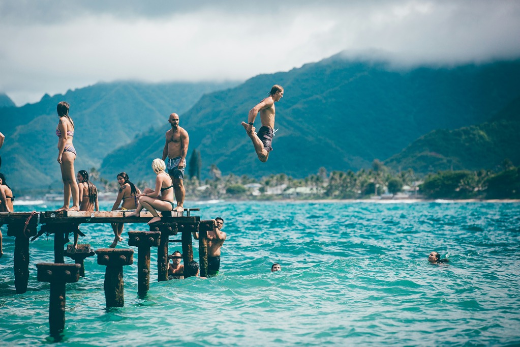A group of friends enjoying summer fun in the sun, jumping from a dock into a lake surrounded by mountains.